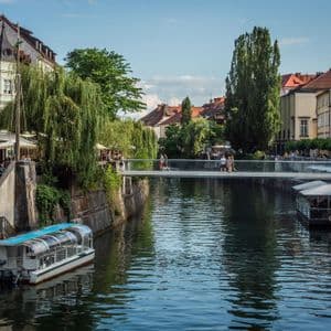 Un moderno puente peatonal de cristal cruza un canal de la ciudad, flanqueado por edificios históricos, cafés ribereños y barcos turísticos.