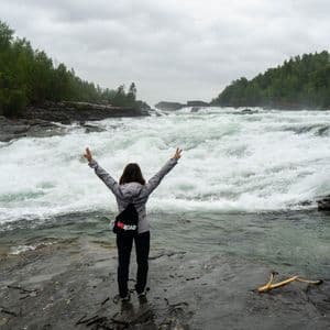 Una persona in viaggio WeRoad si trova su una riva rocciosa con le braccia alzate, di fronte a un fiume turbolento con rapide di acqua bianca.
