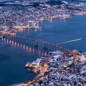 Una vista aerea di una città costiera innevata al tramonto, con un lungo ponte illuminato che attraversa l'acqua verso montagne innevate.