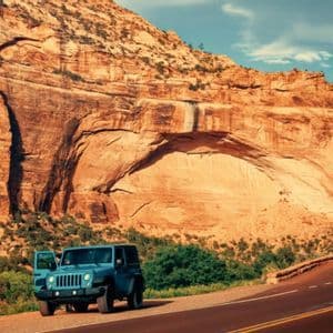 Una jeep blu con la portiera aperta è parcheggiata a bordo strada accanto a una grande formazione rocciosa di canyon rosso.
