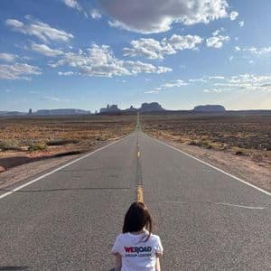 Un capogruppo WeRoad è seduto su una lunga strada dritta che conduce a formazioni rocciose in un paesaggio desertico sotto un cielo blu.