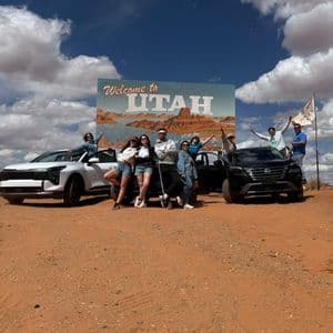 Un gruppo WeRoad posa per una foto con due auto davanti a un grande cartello 'Welcome to Utah' in un paesaggio desertico.