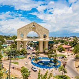 Una veduta aerea di un grande monumento ad arco sopra una rotonda stradale in una città costiera con alberi rigogliosi sotto un cielo azzurro nuvoloso.