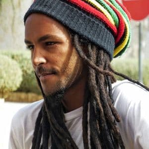 Un uomo con lunghi dreadlocks indossa un berretto a righe rosso, giallo e verde, mentre guarda di lato all'aperto.