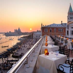 Set tables on a rooftop restaurant overlook a historic city's waterfront with boats and gondolas at sunset.