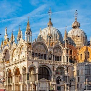 The detailed facade of an ornate basilica with multiple domes and spires stands against a bright blue sky.