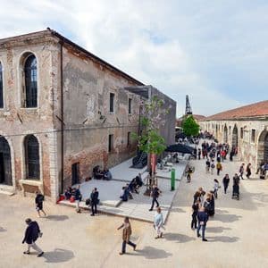 A high-angle shot of people walking and relaxing in a large courtyard between two old, weathered brick buildings under a cloudy sky.
