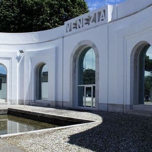 A modern white building with a curved facade, arched windows, and the word 'VENEZIA' carved above, next to a gravel courtyard.