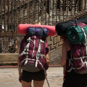 Due persone di un viaggio di gruppo WeRoad, viste di spalle, con grandi zaini e materassini, su una strada di ciottoli.