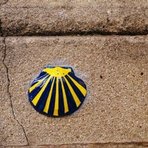 Un primo piano di una conchiglia di capasanta in ceramica blu e gialla, simbolo del Cammino di Santiago, incastonata in un muro di pietra texturizzato.