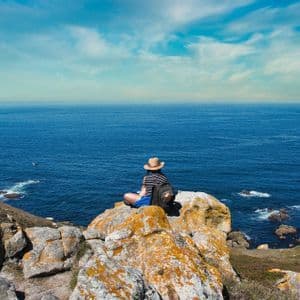 Una persona con un cappello di paglia e uno zaino siede su una scogliera rocciosa affacciata sull'oceano blu.