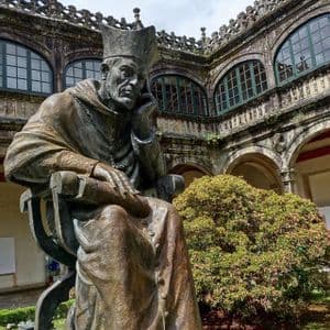 Una grande statua in bronzo di un chierico seduto in posa pensierosa, situata in un cortile storico in pietra con finestre ad arco.