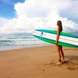 Una donna con la tavola da surf in piedi su una spiaggia di sabbia osserva le onde dell'oceano sotto un cielo parzialmente nuvoloso.
