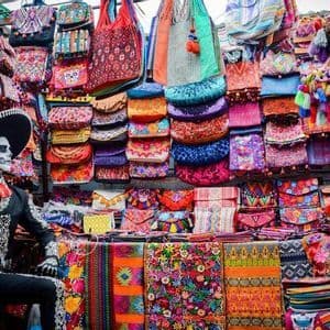 Un manichino in costume del Giorno dei Morti è seduto a una bancarella del mercato, ricca di borse, borsette e tessuti colorati.