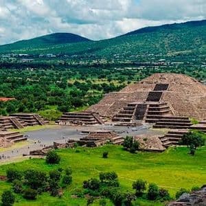 Un'ampia veduta di un antico complesso piramidale, immerso in una lussureggiante valle verde, con dolci colline sullo sfondo sotto un cielo nuvoloso.