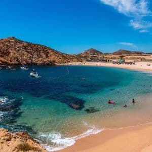 Una caletta soleggiata con una spiaggia sabbiosa, acqua cristallina color turchese, nuotatori e diverse barche ancorate nella baia.