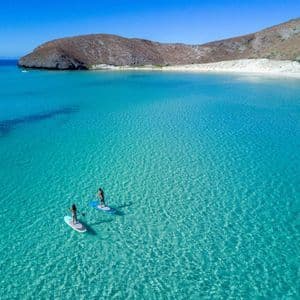 Veduta aerea di due persone su paddleboard in una baia con acque cristalline turchesi e una spiaggia di sabbia bianca.