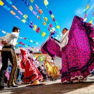 Ballerini in vivaci costumi tradizionali si esibiscono su un palco all'aperto sotto bandiere colorate di festival contro un cielo blu.