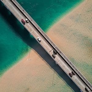 Vue aérienne de voitures roulant sur un pont enjambant des eaux turquoise et un littoral sablonneux.