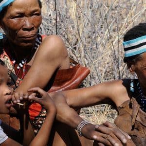 Zwei indigene Frauen und ein kleines Kind sitzen draußen zusammen und tragen traditionelle Kleidung und Perlenschmuck.