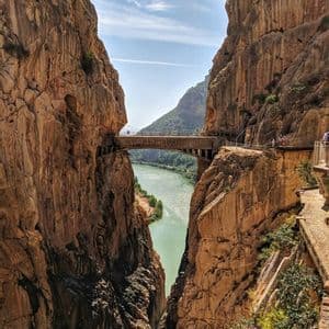 Una pasarela estrecha y un puente adheridos a las escarpadas paredes del cañón, con un río turquesa visible muy abajo bajo un cielo azul.