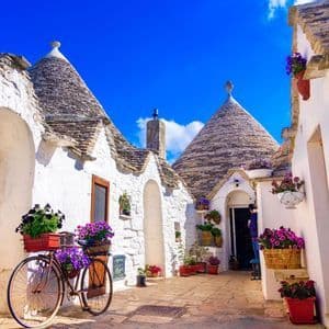 A vintage bicycle with baskets of purple flowers is parked on a stone alley between whitewashed houses with conical roofs under a blue sky.