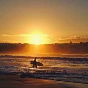 Une personne en silhouette avec une planche de surf marche dans l'océan au coucher du soleil, qui inonde l'eau d'une lueur dorée.