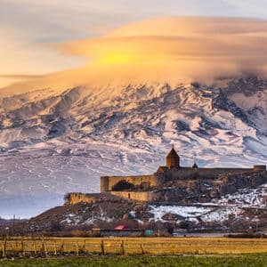Ein steinernes Kloster thront auf einem Hügel vor einem massiven, schneebedeckten Berg, der von einer großen, leuchtenden Wolke gekrönt wird.