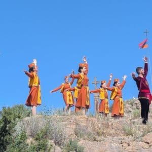 Eine Gruppe von Personen in traditionellen orange-gelben Kostümen vollführt einen synchronisierten Tanz auf einem felsigen Berghang unter strahlend blauem Himmel, neben einer armenischen Flagge.