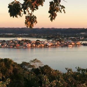 Eine Vogelperspektive einer farbenfrohen Inselstadt an einem ruhigen See bei Sonnenuntergang, umrahmt von Baumblättern im Vordergrund.