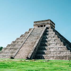 Eine große steinerne Stufenpyramide mit einer zentralen Treppe steht auf einem grünen Rasen unter einem klaren blauen Himmel.