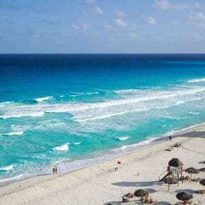Luftaufnahme eines weißen Sandstrandes mit türkisfarbenen Wellen, gesprenkelt mit Strohschirmen und sonnenbadenden Menschen.