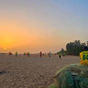 Eine Person in einem gelben Trikot sitzt bei Sonnenuntergang auf Fischernetzen und beobachtet Menschen beim Fußballspielen am Sandstrand.