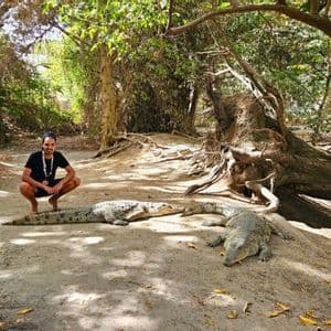 Ein lächelnder Mann hockt barfuß auf einem Sandweg neben drei großen Krokodilen, mit einem üppig grünen Wald im Hintergrund.