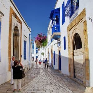 Persone camminano lungo una stretta strada acciottolata, fiancheggiata da edifici bianchi con porte e finestre blu brillante, sotto un cielo sereno.