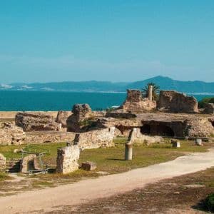 Antiche rovine in pietra su un appezzamento erboso in riva al mare, con montagne lontane visibili oltre l'acqua sotto un cielo sereno.