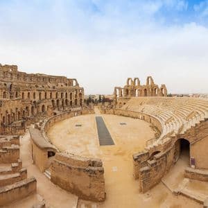 Una vista grandangolare delle rovine di un grande e antico anfiteatro con posti a sedere in pietra a gradoni e un'arena centrale sabbiosa.