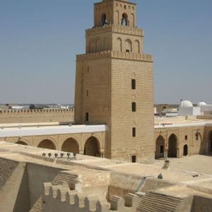 Un minarete cuadrado de piedra color arena con una cúpula se alza sobre los tejados de un antiguo complejo bajo un cielo azul claro.