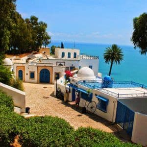 Un borgo di edifici bianchi con porte e finiture blu su una strada di ciottoli, arroccato su una scogliera a picco sull'oceano.