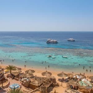 Una vista elevada de una playa de arena con sombrillas de paja, personas nadando en aguas turquesas y varios yates amarrados en el mar.