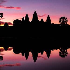 Silhouette di un complesso di templi e palme riflesse in acqua calma durante un tramonto rosa e viola.