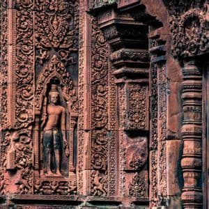 Close-up of intricate carvings on a reddish stone temple wall, featuring a standing deity in a decorative niche.