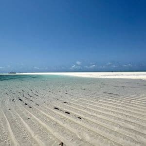Rippled sand patterns in shallow, clear water on a white sand beach under a vast, cloudless blue sky.