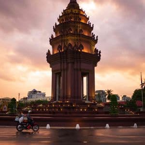 Un monument orné et illuminé se dresse sur une place de ville au coucher du soleil, tandis que deux motos passent sur une route mouillée.