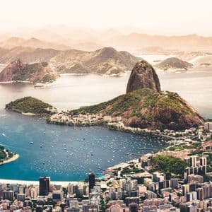 An aerial view of a sprawling coastal city with a large mountain overlooking a bay filled with boats, with hazy hills in the distance.