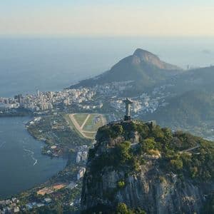 An aerial view of the Christ the Redeemer statue on a mountain, overlooking a coastal city with a large lagoon and the ocean.