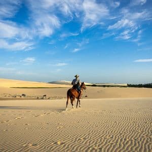 Una persona con un cappello cavalca un cavallo marrone attraverso vaste dune di sabbia ondulate sotto un cielo azzurro brillante con nuvole sottili.