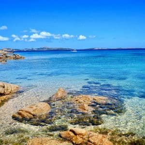 Una costa rocciosa incontra acque calme, limpide e turchesi sotto un cielo azzurro brillante con una singola barca in lontananza.