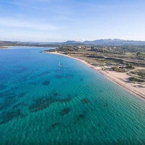 Vista aerea di una barca a vela solitaria su acqua cristallina turchese lungo una spiaggia sabbiosa, con colline e montagne verdi in lontananza.