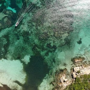 Veduta aerea di una piccola barca che naviga su acque turchesi limpide, lasciando una scia, vicino a una costa rocciosa.